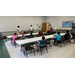 A group of children sitting at tables while listening to a woman speak.