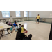 Children sitting down in the back of a classroom with a teacher at the front of the room.